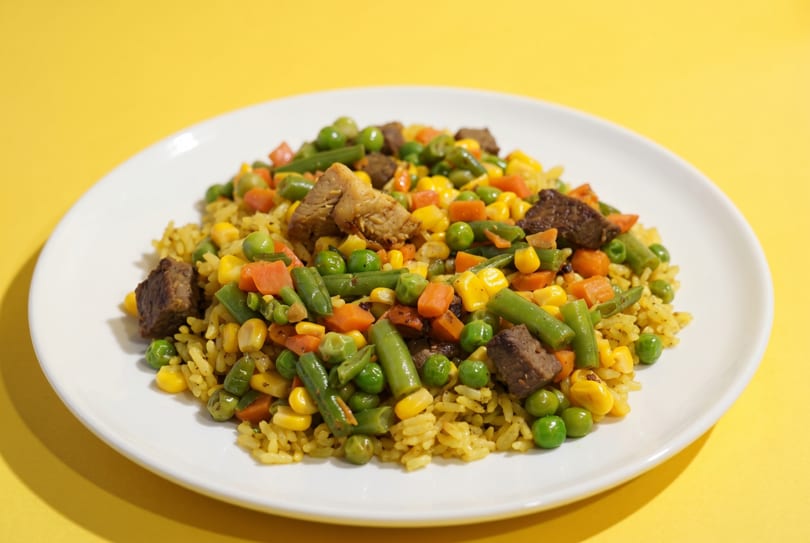 A white plate holds yellow rice mixed with green beans, peas, corn, carrots, and beef chunks.